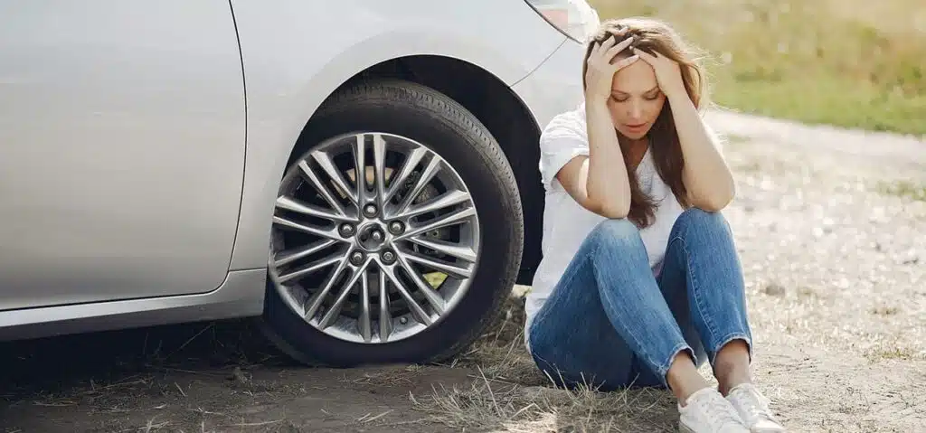 woman with both hands on her head seating beside a car looking traumatized